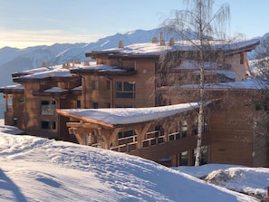 A multi-story building with a wooden facade and snow-covered roofs, set against a mountainous backdrop. - Spacious apartment, mountain view - 5 guests (Les Arcs 1800)