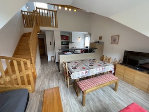 A loft-style living space with a wooden staircase, a dining area with a table and bench, a kitchen with modern appliances, and a television on a wooden cabinet. - Duplex apartment with parking - 6 guests (Le Lioran)