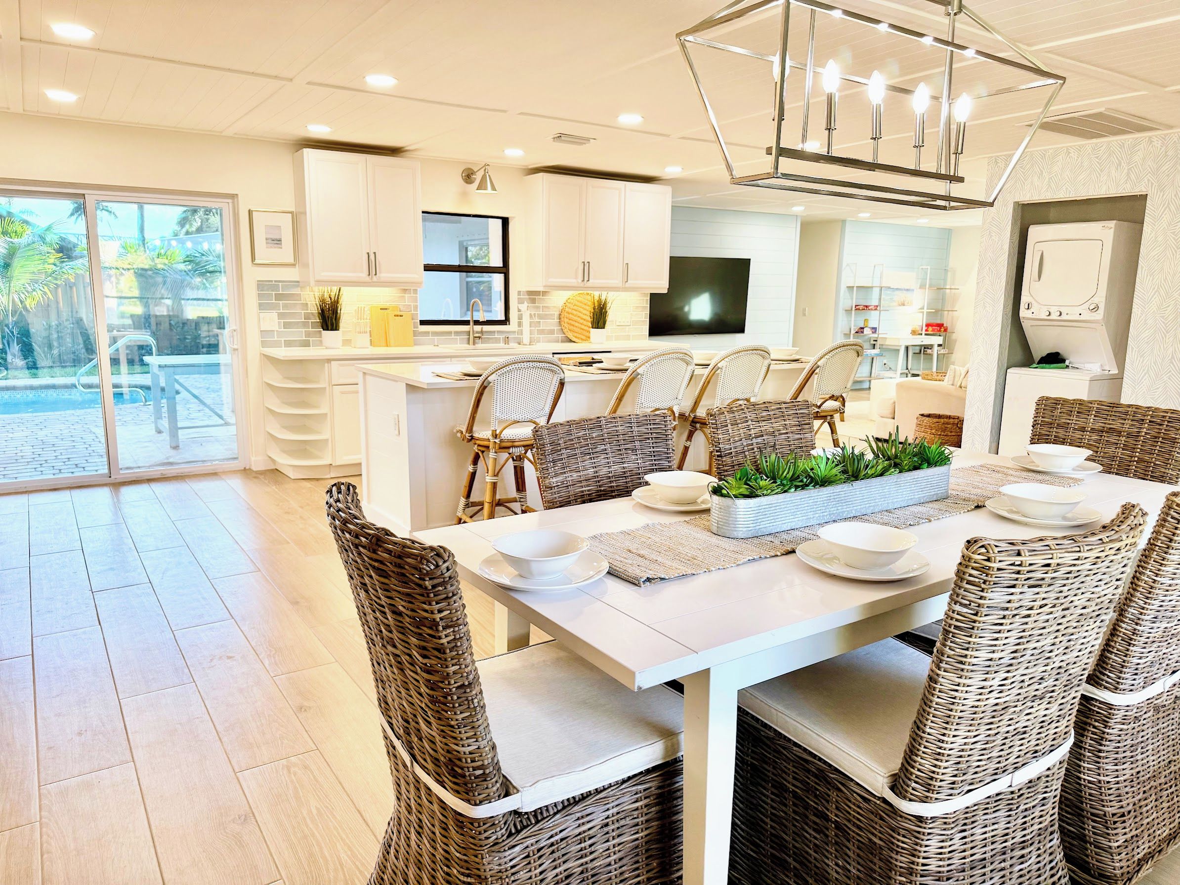 A modern kitchen with white cabinets, a dining area with wicker chairs, and a large window overlooking a garden.