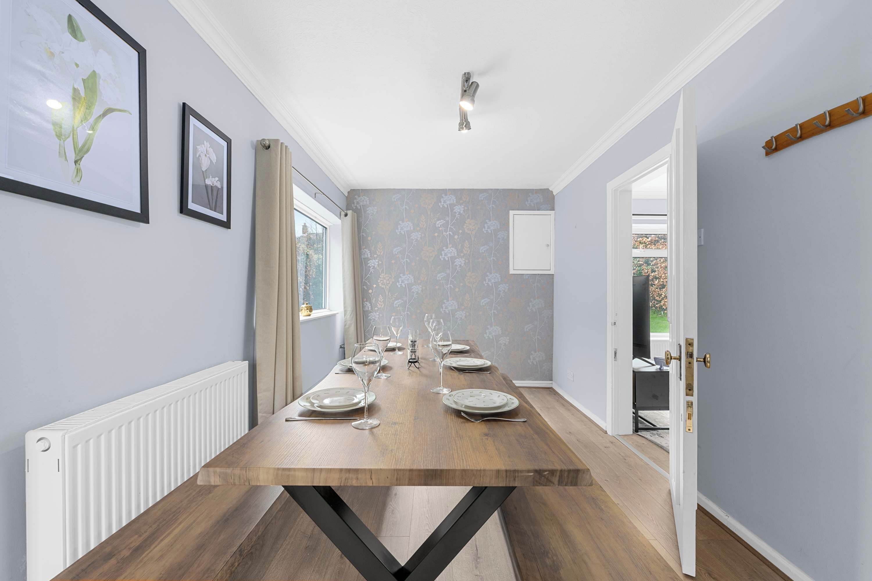 A dining area with a long wooden table set for six, flanked by light-colored walls and a window.