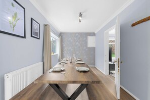 A dining area with a long wooden table set for six, flanked by light-colored walls and a window. - Hale Barns Home - Garden, Parking & Near Airport (Hale)