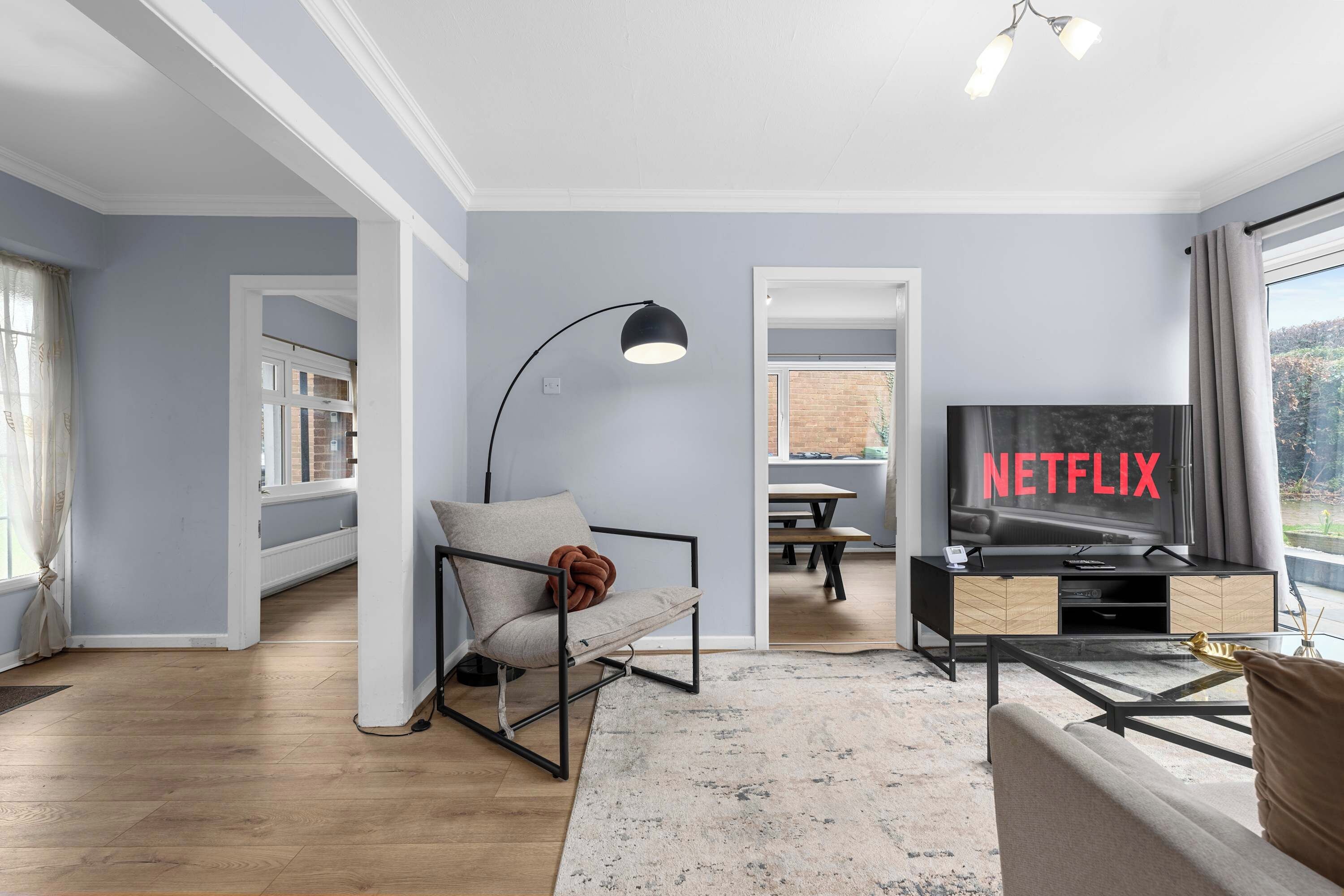 A modern living room with a sofa, a black floor lamp, a TV displaying 'NETFLIX', and a dining area in the background.