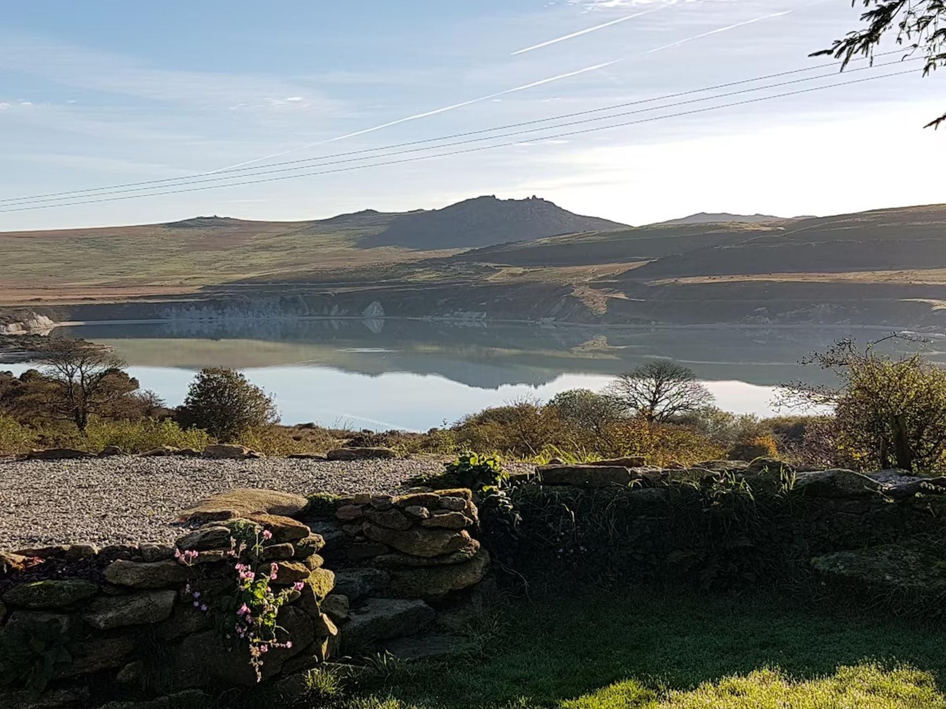 A tranquil lake with a stone wall and vegetation in the foreground, surrounded by hills and a clear sky.