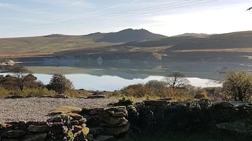 Ein ruhiger See mit einer Steinmauer und Vegetation im Vordergrund, umgeben von Hügeln und einem klaren Himmel.