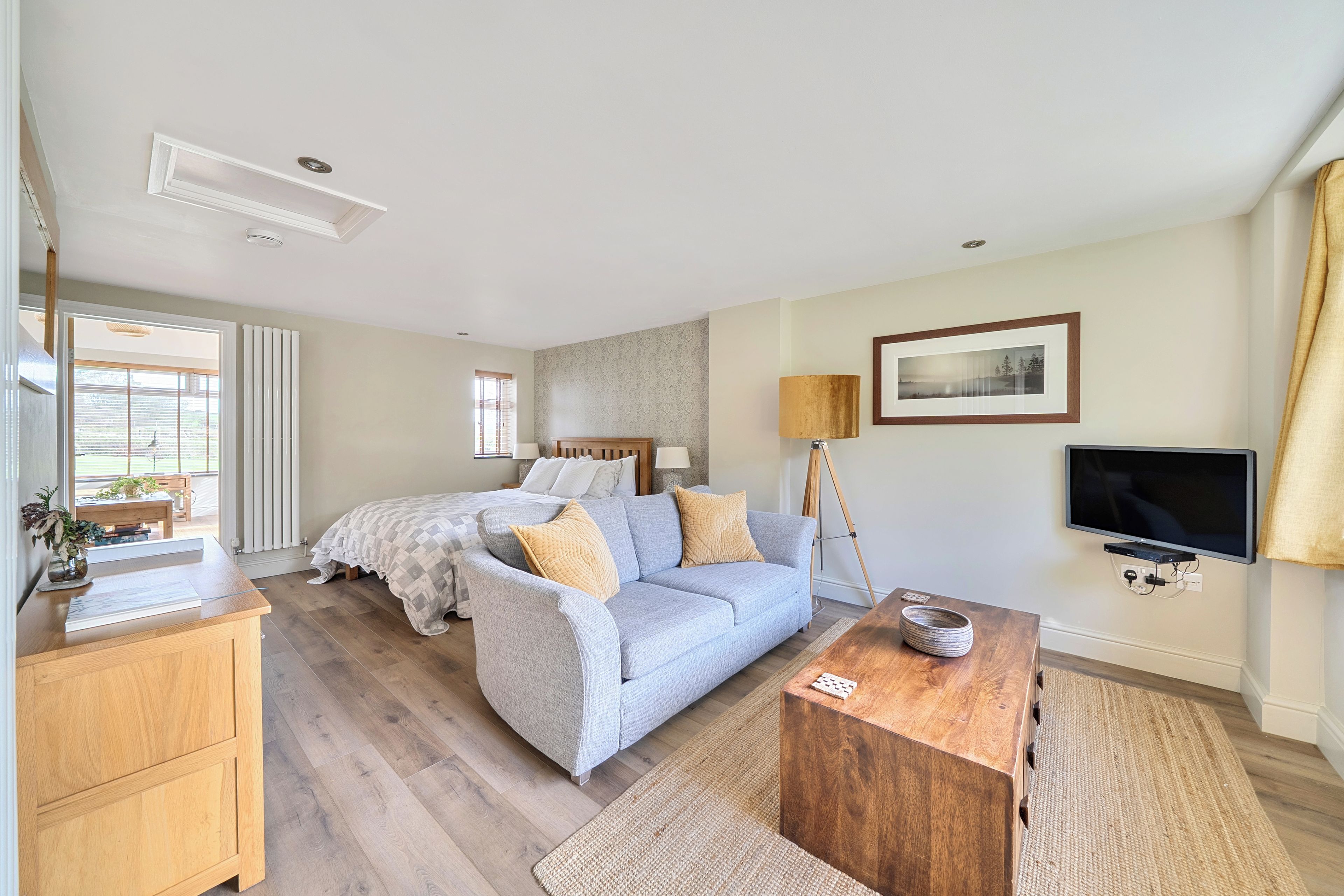 A spacious bedroom with a large bed, a sofa, a wooden side table, a television, and a window with blinds.