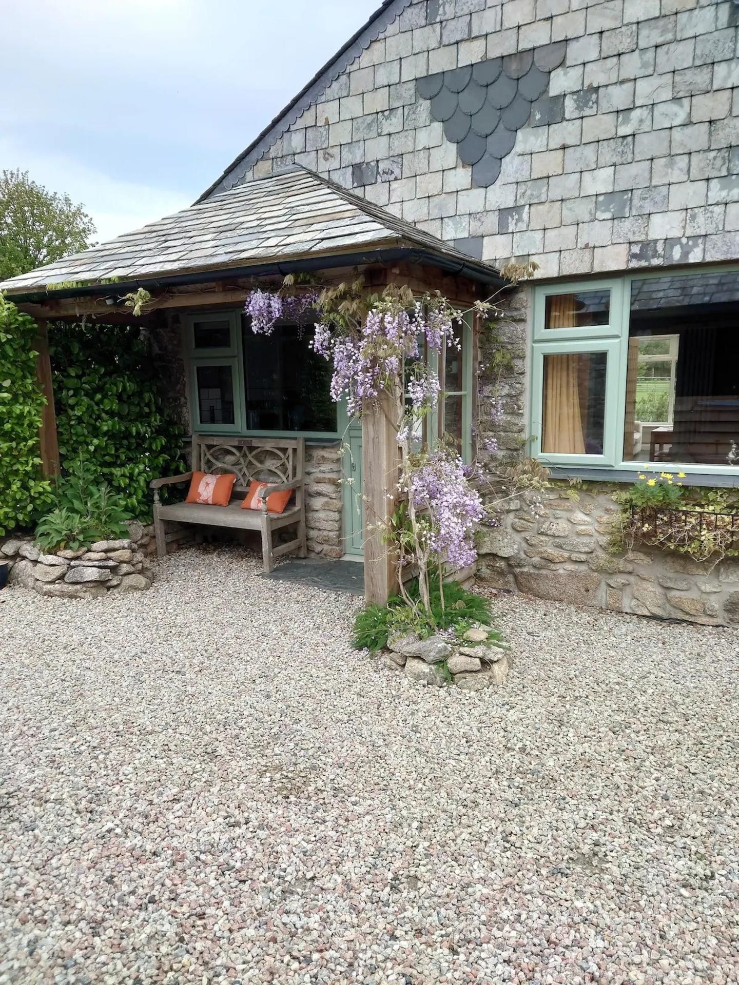 A stone house with a gravel driveway, a wooden bench, and purple flowers.