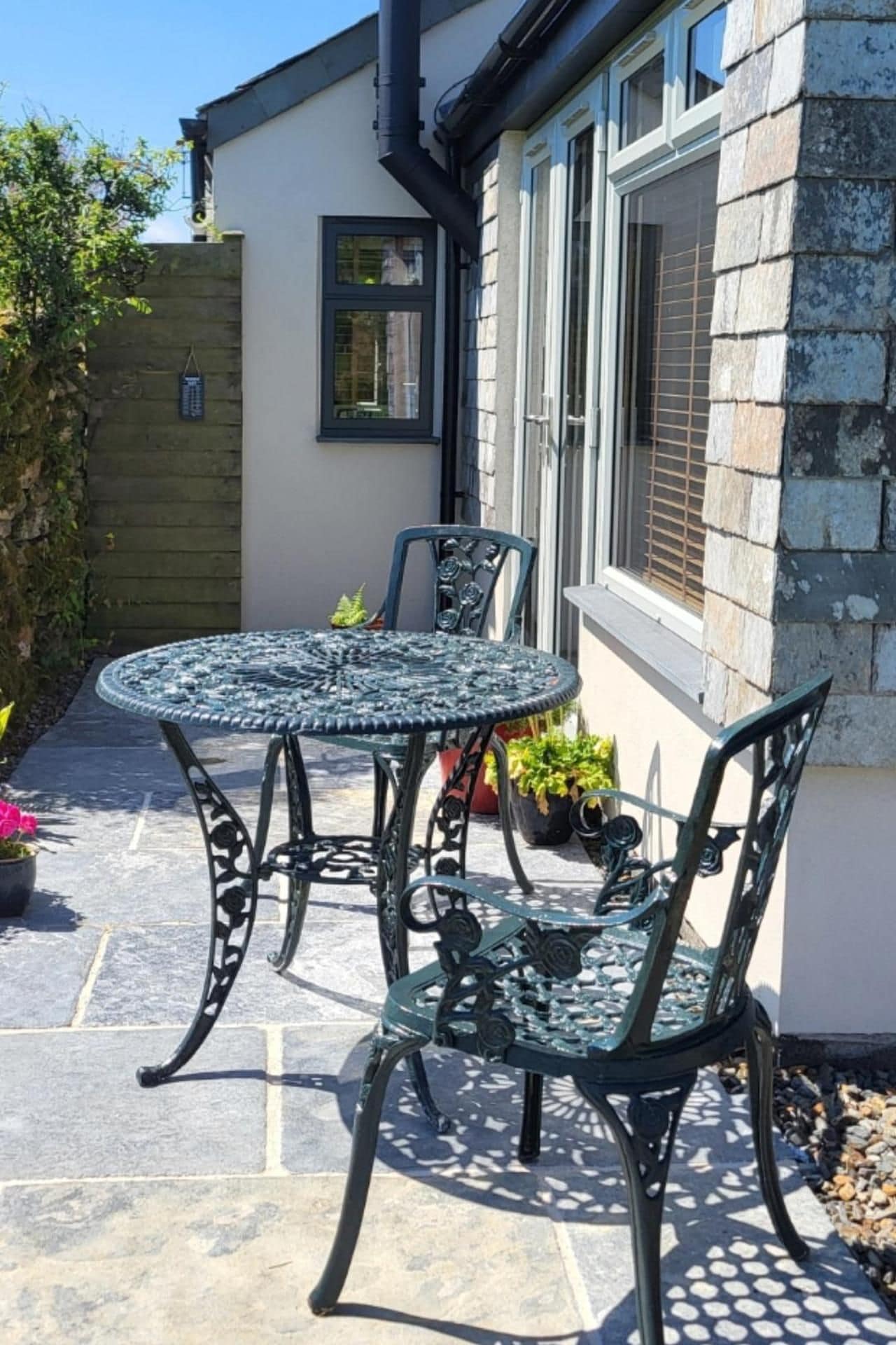 A patio with a round table and chairs, a window with blinds, and a stone wall.