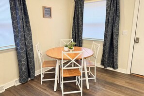 Un coin repas avec une table ronde en bois et quatre chaises blanches, une plante en pot posée sur la table et des rideaux à motifs aux fenêtres.