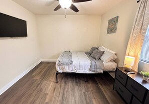 Een slaapkamer met een bed, een plafondventilator, een aan de muur bevestigde televisie, een bedlampje en een raam met gordijnen.