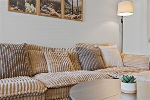 Uma sala de estar moderna com um sofá, vários almofados, uma mesa de centro redonda, um abajur e uma pequena planta em vaso.