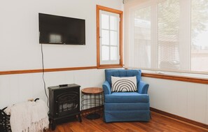 Ein Zimmer mit an der Wand montiertem Fernseher, ein blauer Sessel mit gemustertem Kissen, ein Holzboden und ein kleiner runder Tisch mit Drahtgestell.