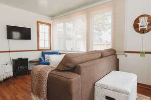 A cozy living room with a brown sofa, a blue armchair, a white ottoman, a television, and a wooden chair on the wall. - The Driftwood Cottage - Lake Huron Access (Lambton Shores)