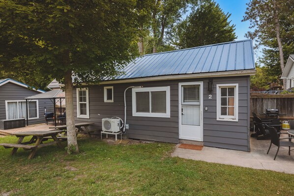 En liten, envåningsstuga med ett tak av metall, en ytterdörr och en liten veranda med ett picknickbord och stolar.