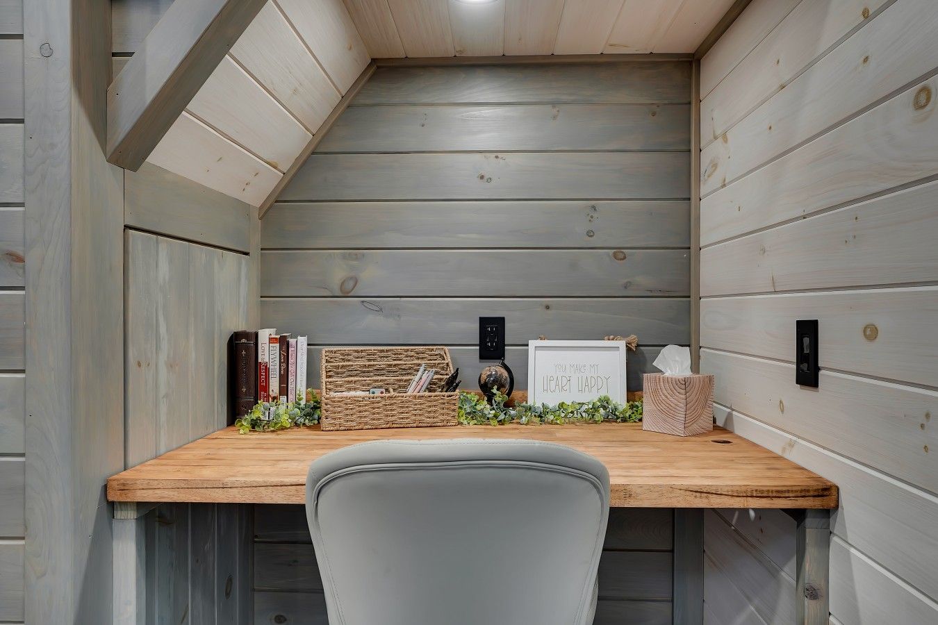 Een hoekzaal met een houten bureau, een grijze stoel, boeken, een lijst met een afbeelding en een kleine plant.