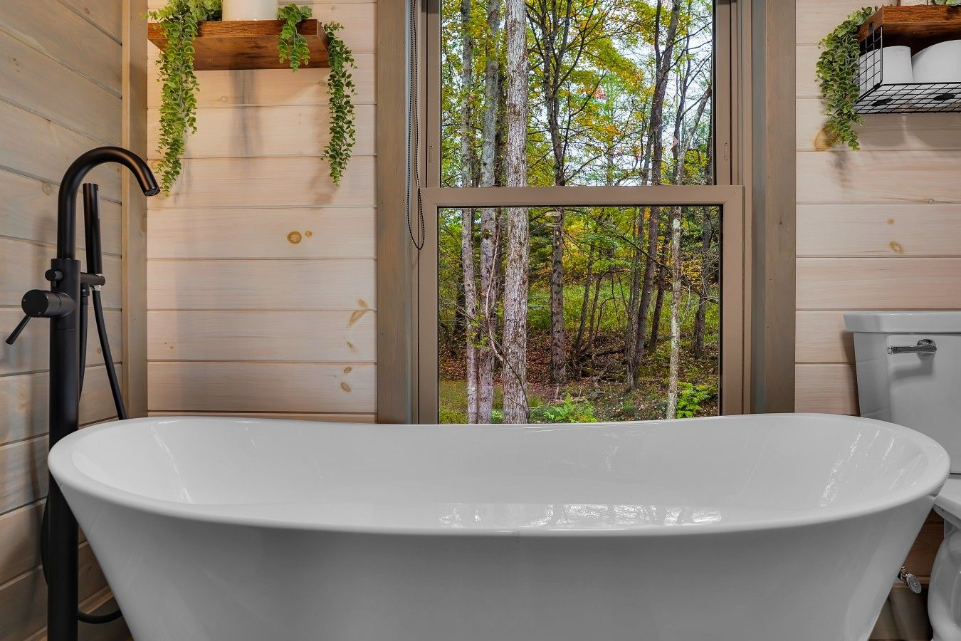 A bathroom with a freestanding bathtub, a toilet, and a window with a view of a forest.