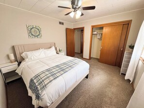 Un dormitorio con cama, ventilador de techo, mesita de noche, puerta y piso alfombrado.