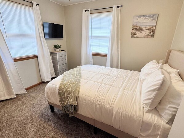 A bedroom with a bed, curtains, a flat-screen TV, a small dresser, and a painting on the wall.