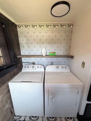 A compact laundry room with two white front-loading washers and dryers, a small shelf above the dryers, and a patterned wall. - The Farm House at Sunnybrook Farms (Van Buren)