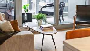 A modern living room with a sofa, a coffee table with books and a plant, and a view of the outside through large windows. - Melbourne Skyline High-Rise Suite | City Views & Pool Access (Melbourne)