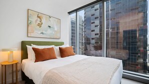 A hotel room with a large bed, a green upholstered headboard, a white throw blanket, and a view of tall buildings through a large window. - Melbourne Skyline High-Rise Suite | City Views & Pool Access (Melbourne)