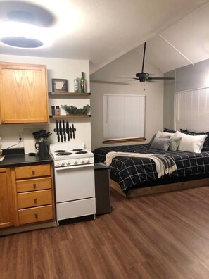 Een kleine kamer met een kitchenette, een bed met zwart-wit geruit beddegoed, een plafondventilator en een raam met jaloezieën.