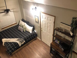 A bedroom with a bed, a door, a shelf with books and glasses, and a ceiling fan.