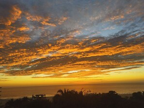 A sunset over a coastal landscape with silhouettes of palm trees and a distant view of the ocean. - Oceanfront Apartment w/ Surf Views & Pool Access (Puerto Escondido)