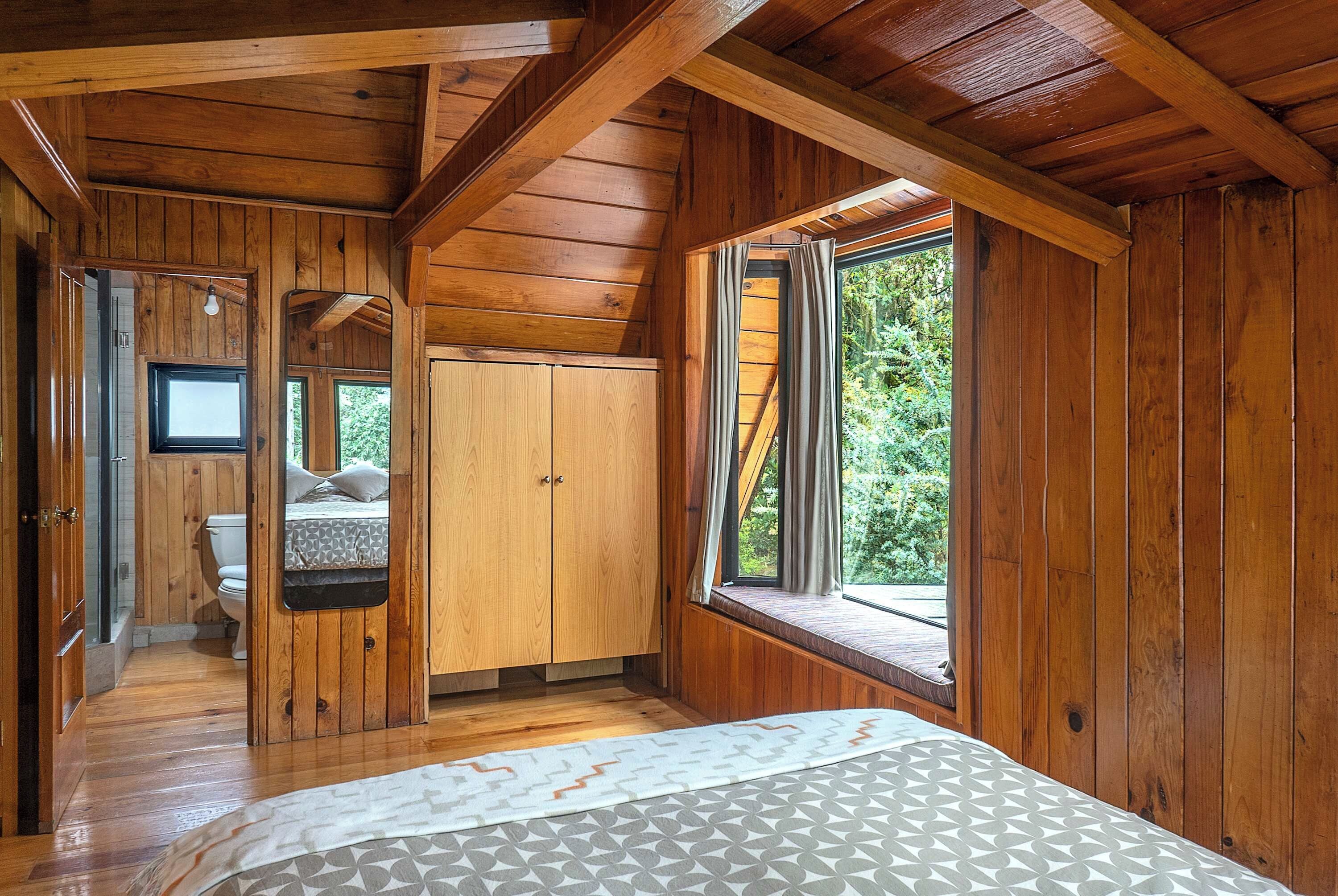 L’intérieur d’un chalet en bois, avec un lit, une fenêtre avec des rideaux, une porte et une vue sur la verdure à l’extérieur.