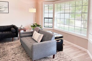 A living room with a grey sofa, a black armchair, a small table with a lamp, and a window with blinds. - Happy Heart House | Hot Tub | Firepit (Eugene)