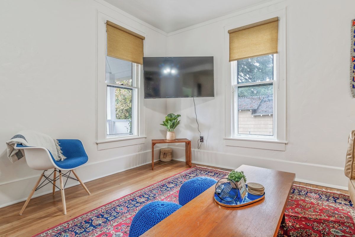 A living room with a wooden coffee table, a blue chair, a TV mounted on the wall, and a window with a view of a house.