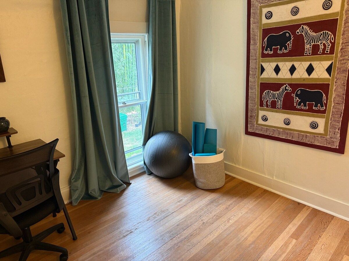 A room with a wooden floor, a desk with a chair, a large exercise ball, and a basket with yoga mats.
