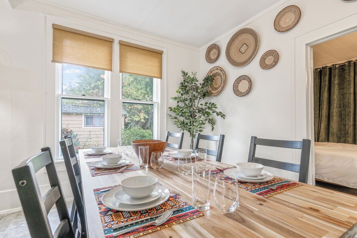 Un comedor con una mesa de madera dispuesta para cuatro personas, rodeada de sillas, con una ventana y persianas a la izquierda, y una cama con cortina a la derecha.
