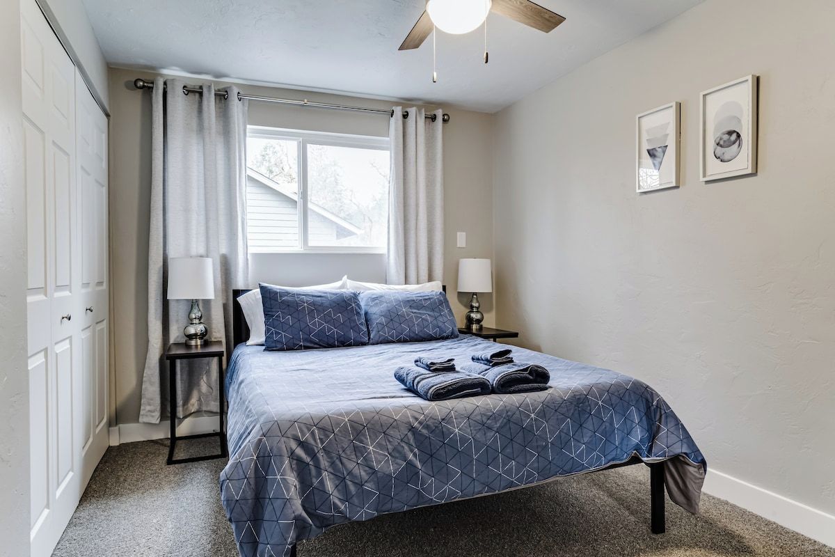 A bedroom with a bed, two bedside tables, a window with curtains, and two framed pictures on the wall.
