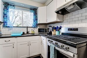 A modern kitchen with white cabinets, a stainless steel stove, a microwave, and a window with patterned curtains. - PNW Forest Getaway w/Fenced Yard & Central Air (Eugene)
