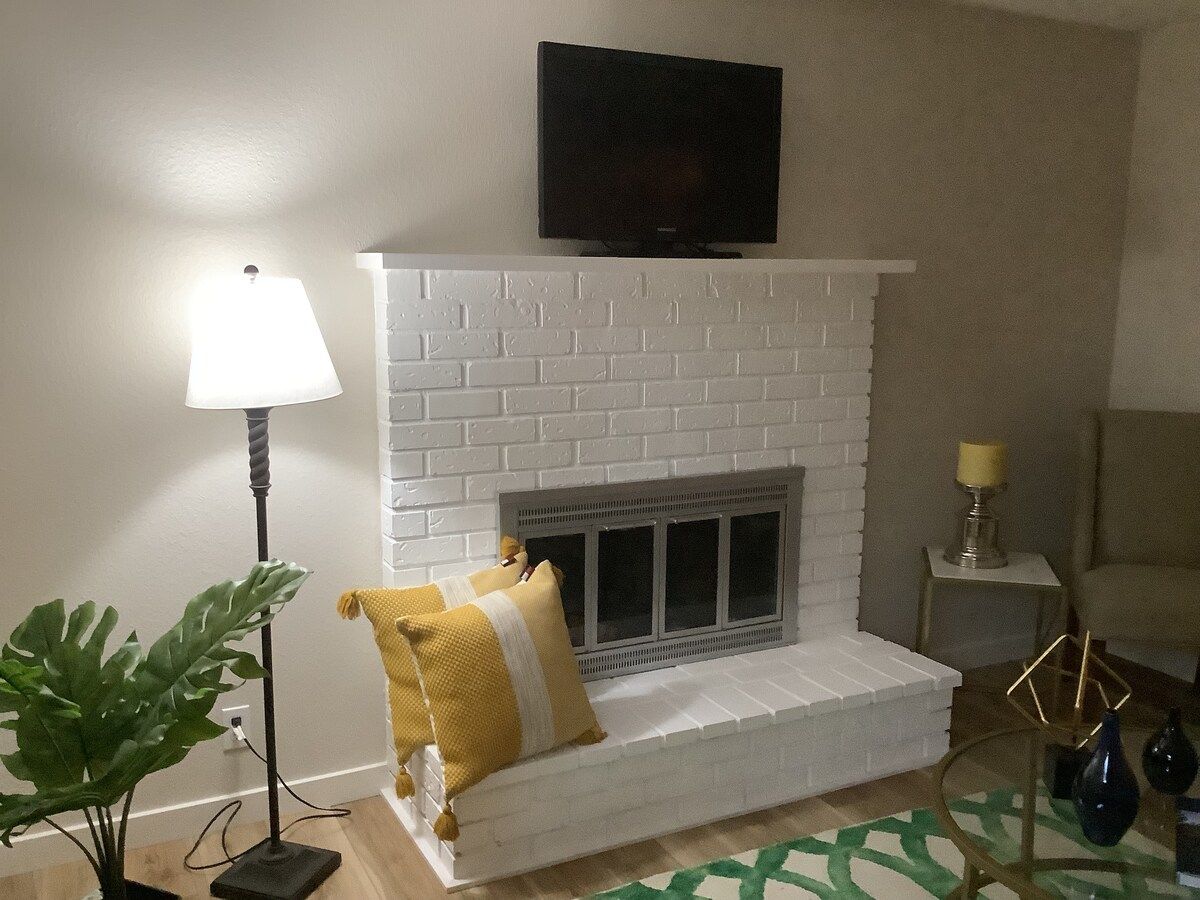 Un salon avec une cheminée en briques blanches, un téléviseur à écran plat fixé au-dessus, un lampadaire, une plante en pot et un tapis à motifs.