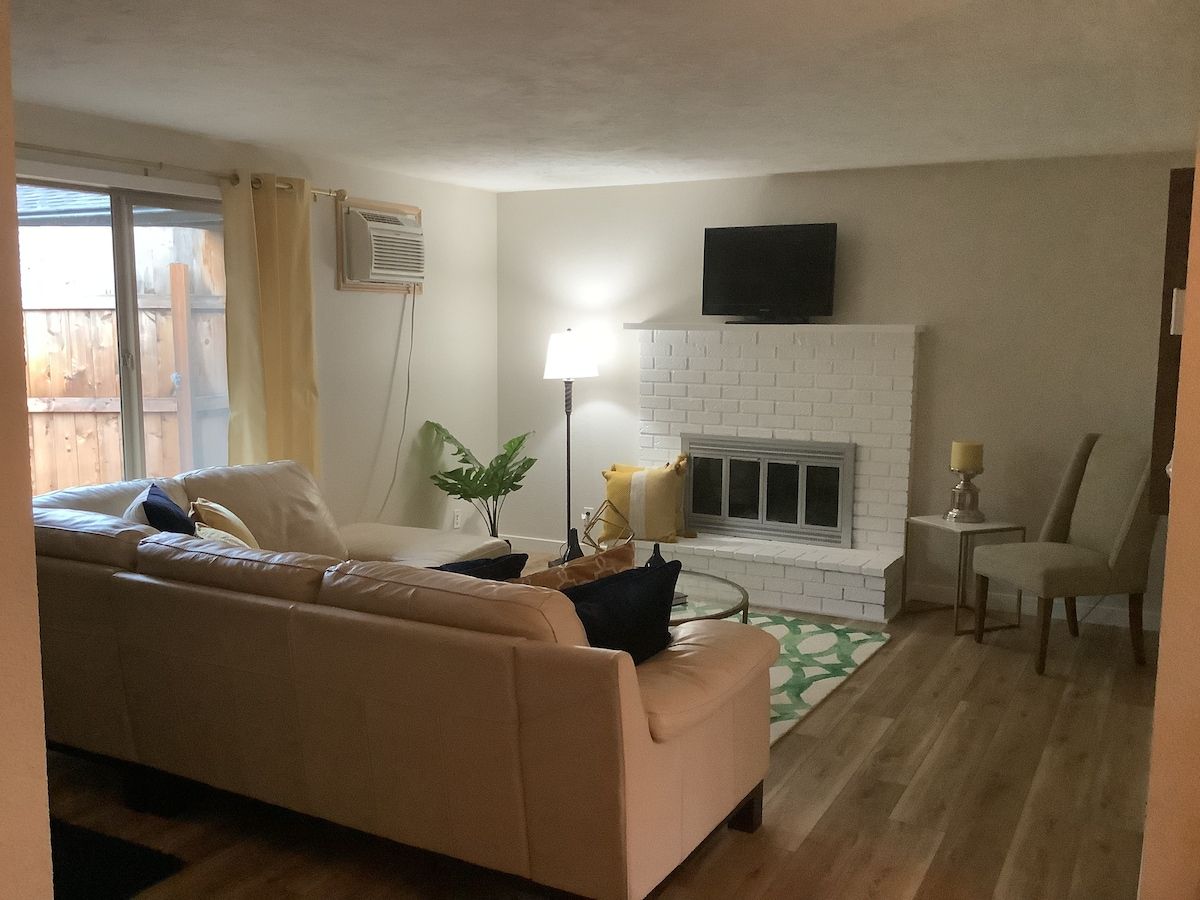 A living room with a beige sofa, a white brick fireplace, a flat-screen TV mounted above it, and a small side table with a lamp.