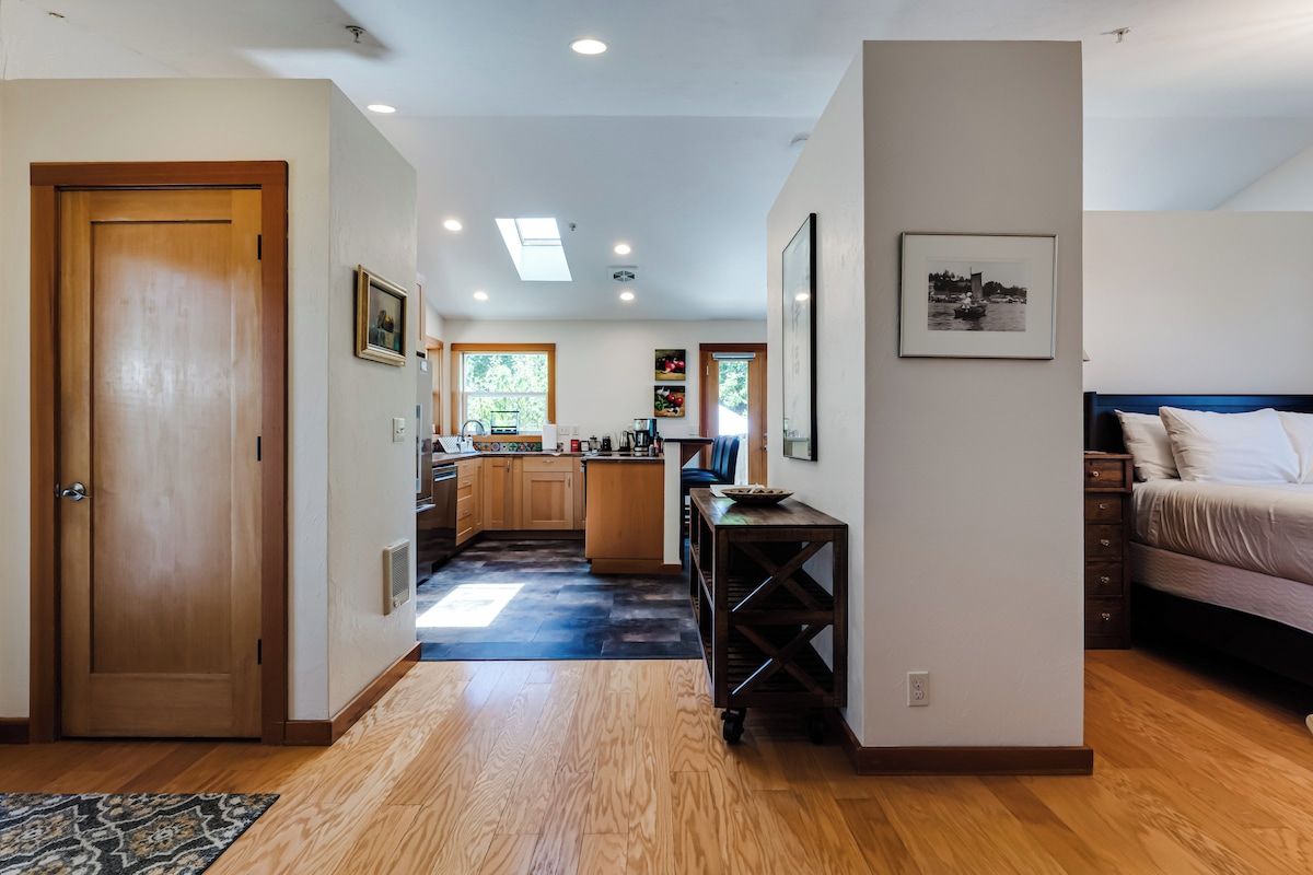 Un intérieur moderne avec un plancher en bois, une cuisine dotée de meubles en bois, un coin repas avec une table et des chaises, et une chambre à coucher avec un lit et une table de chevet.