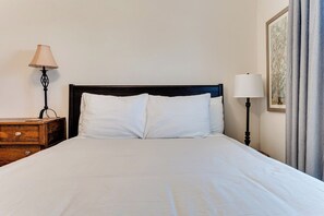 Un lit bien fait, avec du linge de lit blanc, une table de chevet en bois sur laquelle se trouve une lampe, et un tableau encadré accroché au mur.