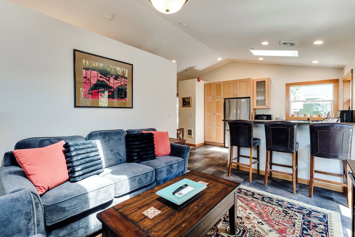 A modern living room with a sofa, coffee table, and a kitchen area with a refrigerator and bar stools.
