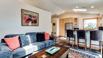 A modern living room with a sofa, coffee table, and a kitchen area with a refrigerator and bar stools.