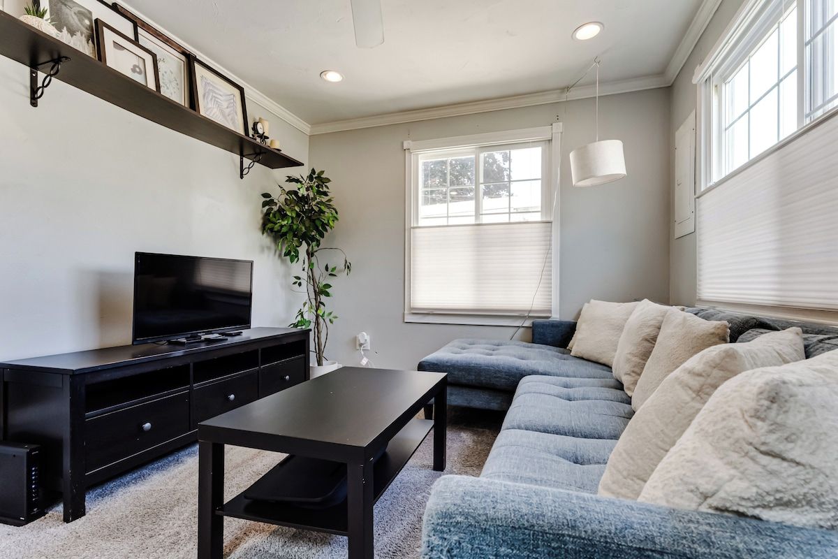 A living room with a black entertainment unit, a grey sofa, and a large window with blinds.
