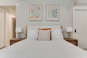 Un dormitorio con una cama, dos mesitas de noche, una lámpara fijada a la pared y dos cuadros enmarcados.