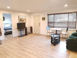 Ein Wohnzimmer mit Flachbildfernseher, Esstisch mit Stühlen, einem grünen Sofa und einem Fenster mit Jalousien.
