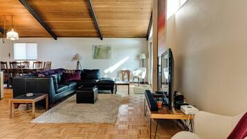 A modern living room with a herringbone floor, a black leather sofa, a white armchair, a wooden coffee table, and a television on a stand.