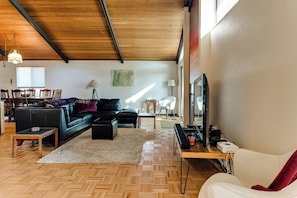 Ein modernes Wohnzimmer mit Fischgrätparkett, einem schwarzen Ledersofa, einem weißen Sessel, einem Holztisch und einem Fernseher auf einem Standfuß.