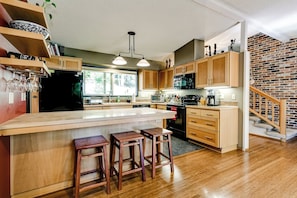A modern kitchen with wooden cabinets, a large island with stools, and a brick wall staircase. - Scenic Eugene Home w/ Deck 1mi to UofO (Eugene)