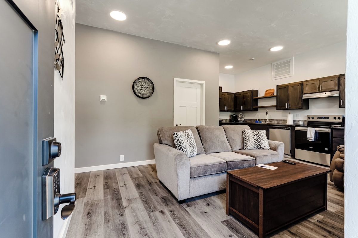 A modern living room with a grey sofa, wooden coffee table, and a kitchen in the background.
