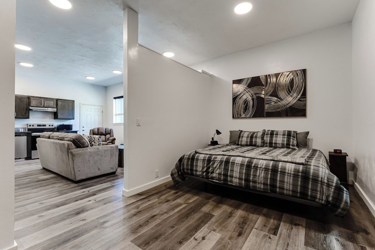 A bedroom with a bed, a couch, and a kitchen in the background.