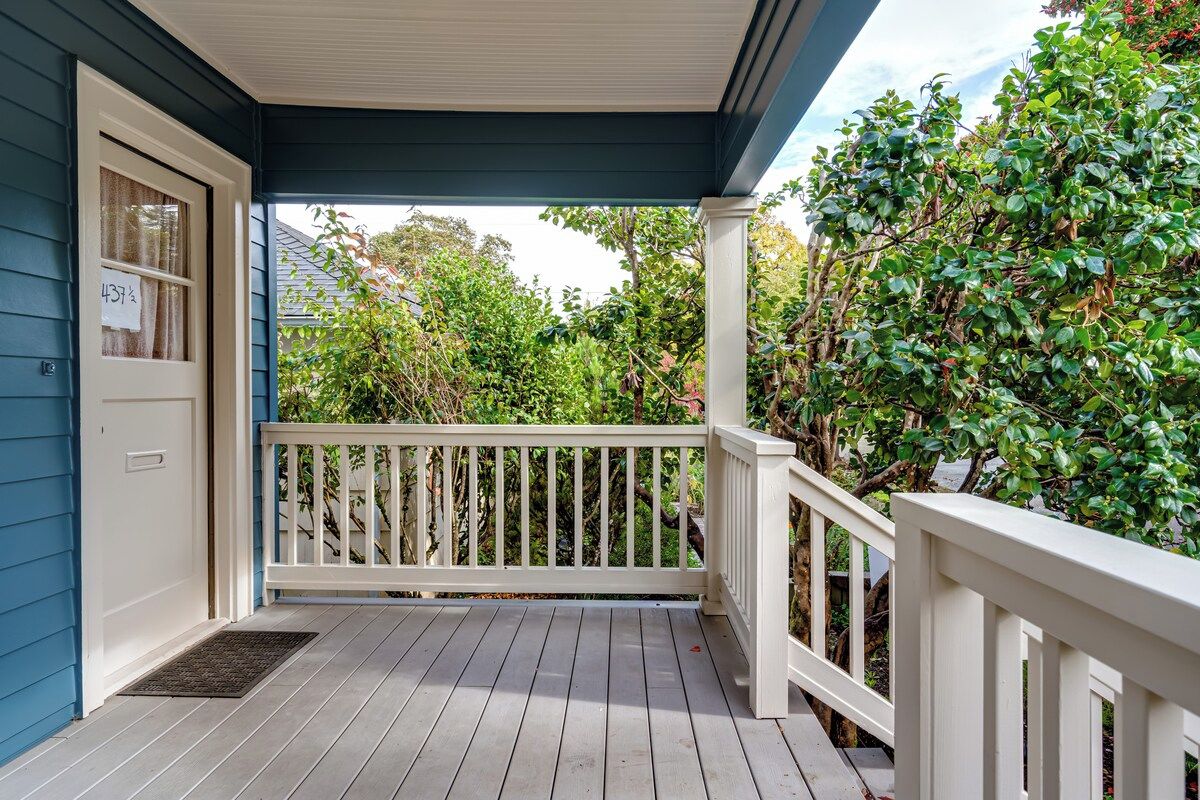 A covered porch with a white railing, a door with a price tag, and a view of greenery.