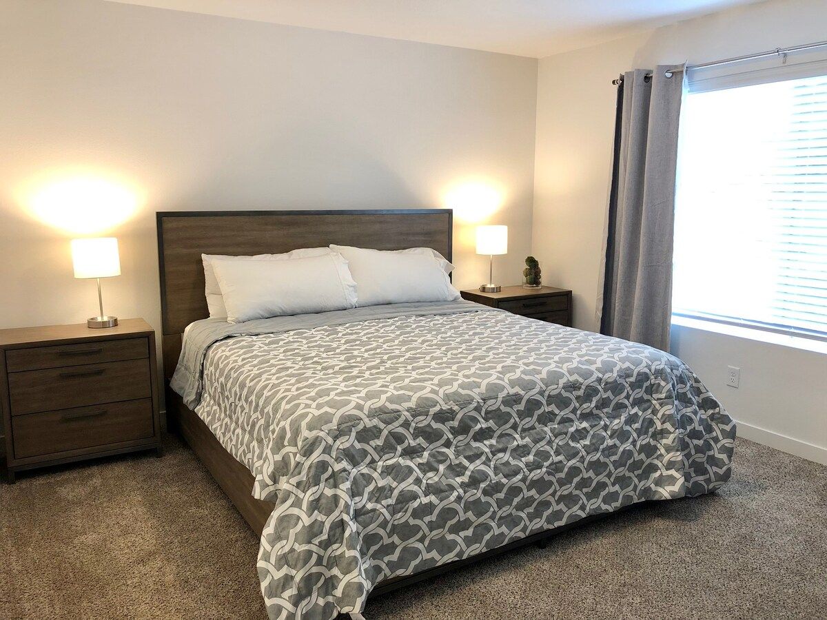 A bedroom with a bed, two bedside tables, a lamp, a window with curtains, and a carpeted floor.